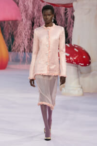 Model on a runway wearing a pale pink textured blazer with pearl buttons and a sheer pink skirt, accessorized with dangling earrings.