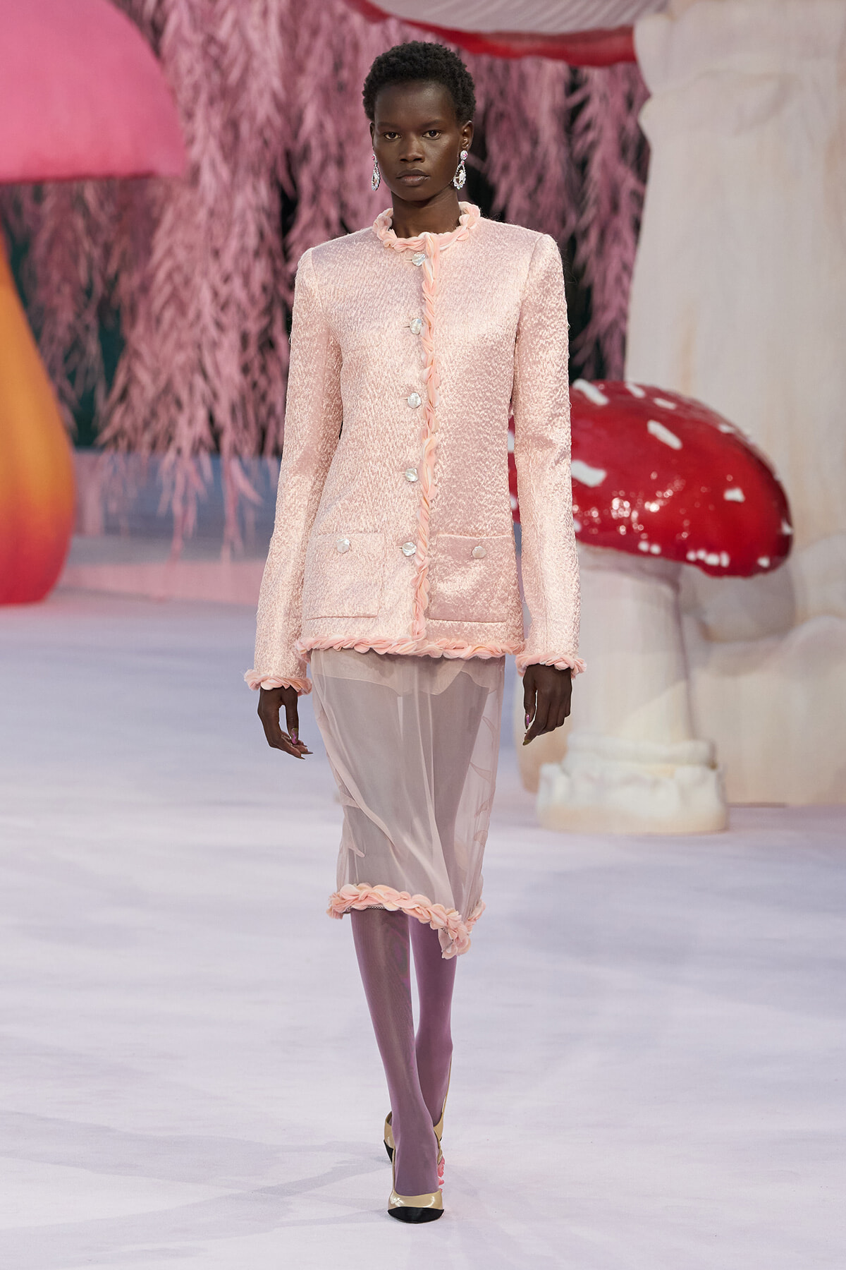 Model on a runway wearing a pale pink textured blazer with pearl buttons and a sheer pink skirt, accessorized with dangling earrings.