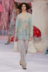 Model walks a fashion runway in a light turquoise sheer embroidered cardigan and matching pants with floral appliqués and pearls.