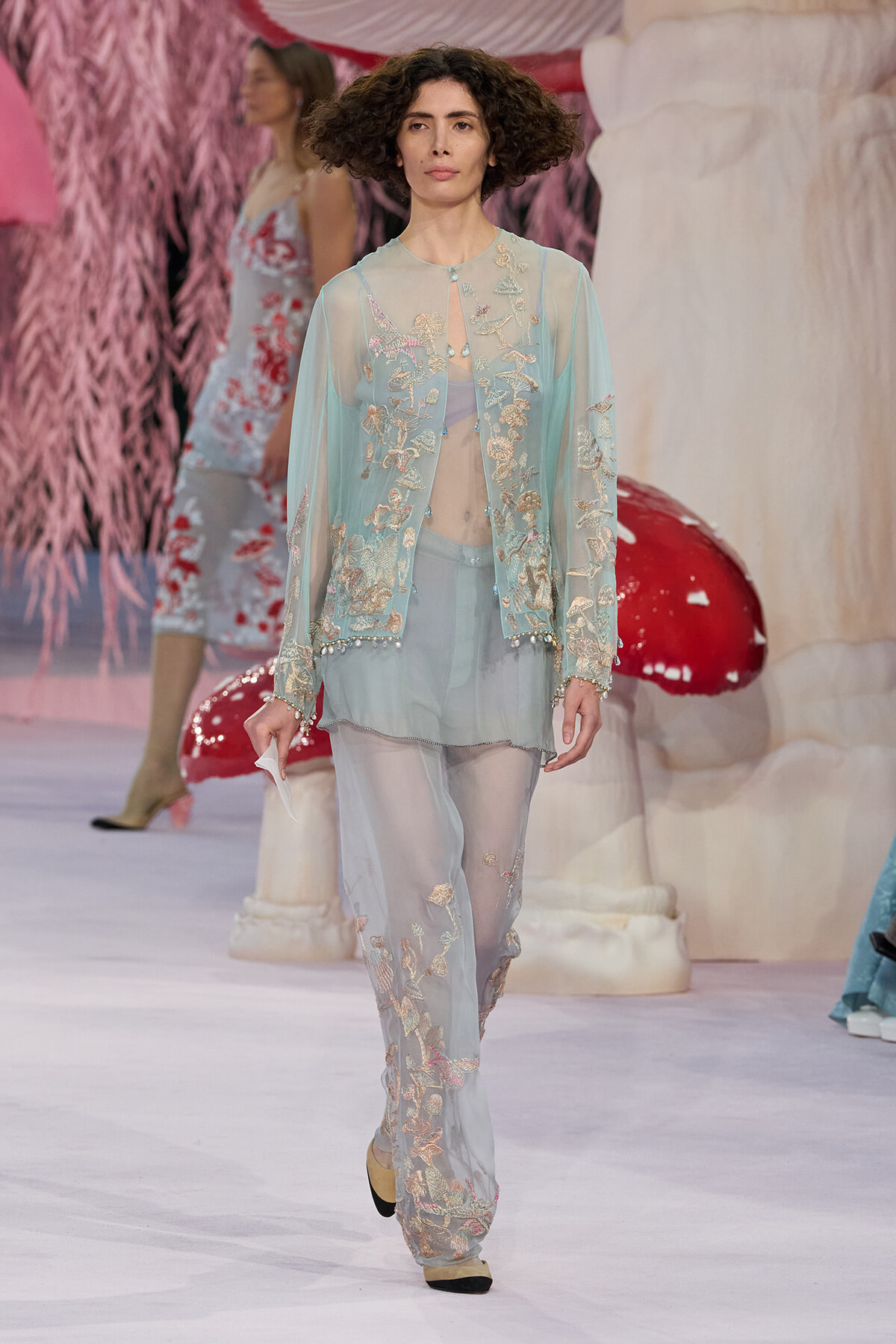 Model walks a fashion runway in a light turquoise sheer embroidered cardigan and matching pants with floral appliqués and pearls.