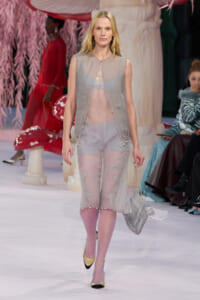 Model walking a fashion show in a sheer gray ensemble: sleeveless vest over a sheer top and scalloped shorts, lavender tights, and two-tone gold-and-black heels, carrying a silver chain-strap bag.