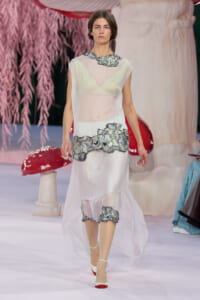 Model on a runway wearing a sheer white dress with grey floral embroidery at the waist and hem, white heels, and pink accents in the background.