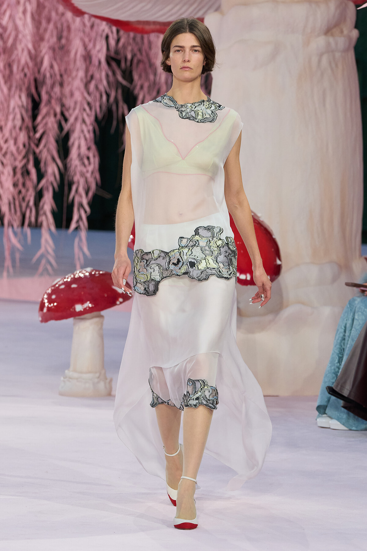 Model on a runway wearing a sheer white dress with grey floral embroidery at the waist and hem, white heels, and pink accents in the background.
