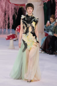 Model walks the runway in a pastel, sculptural gown with layered textures and ruffled black accents amid a fashion show setting.