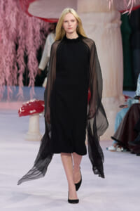Female model walking a runway in a black midi dress with sheer cape sleeves and blonde hair.