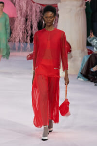Model walks the runway in a sheer red outfit with bead accents, carrying a red handbag.