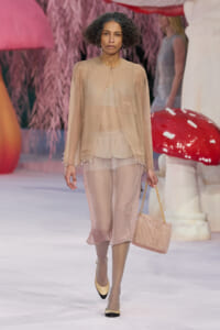 Model walking a runway in a sheer beige two-piece outfit with a matching bag and yellow-black heels amid whimsical pink background scenery.