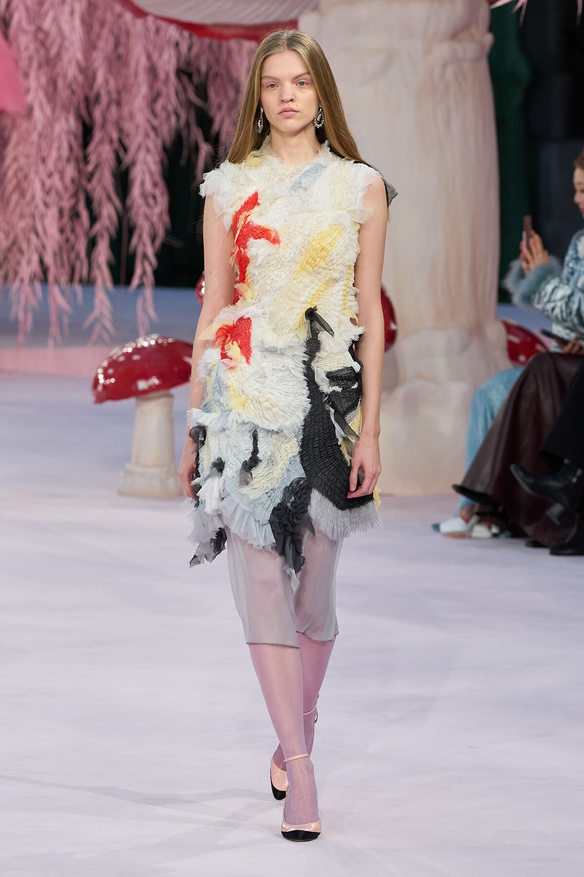 Model walks a fashion runway in a sleeveless, textured cream dress with red, yellow and black appliqués and sheer pink tights.