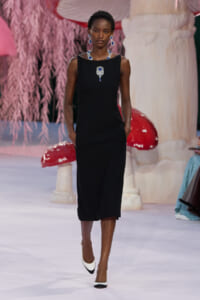 Model walking runway in a sleeveless black midi dress with blue jewelry (necklace and earrings) against a whimsical pink backdrop.