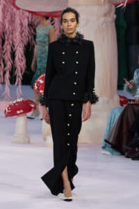 Model walks a runway in a black double-breasted coat with feather-trim cuffs and green buttons, paired with a matching long skirt with a button row.