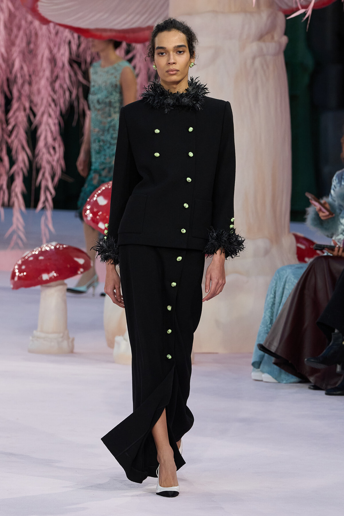 Model walks a runway in a black double-breasted coat with feather-trim cuffs and green buttons, paired with a matching long skirt with a button row.
