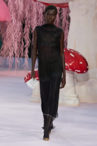 Model walks the runway in a sheer black lace dress, carrying a small handwritten note, gold shoes, with a pink fantasy backdrop behind.