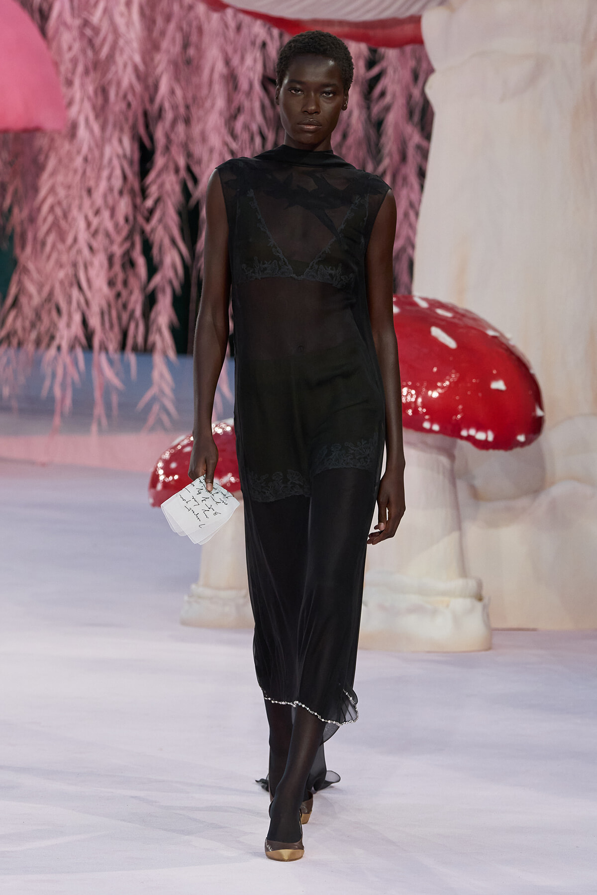 Model walks the runway in a sheer black lace dress, carrying a small handwritten note, gold shoes, with a pink fantasy backdrop behind.