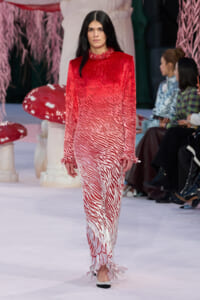 Model walks runway in a long red-to-white zebra-printed dress with ruffled high neck and cuffs. Or concise: Model on runway wearing a gradient red-to-white zebra print gown with ruffle details.