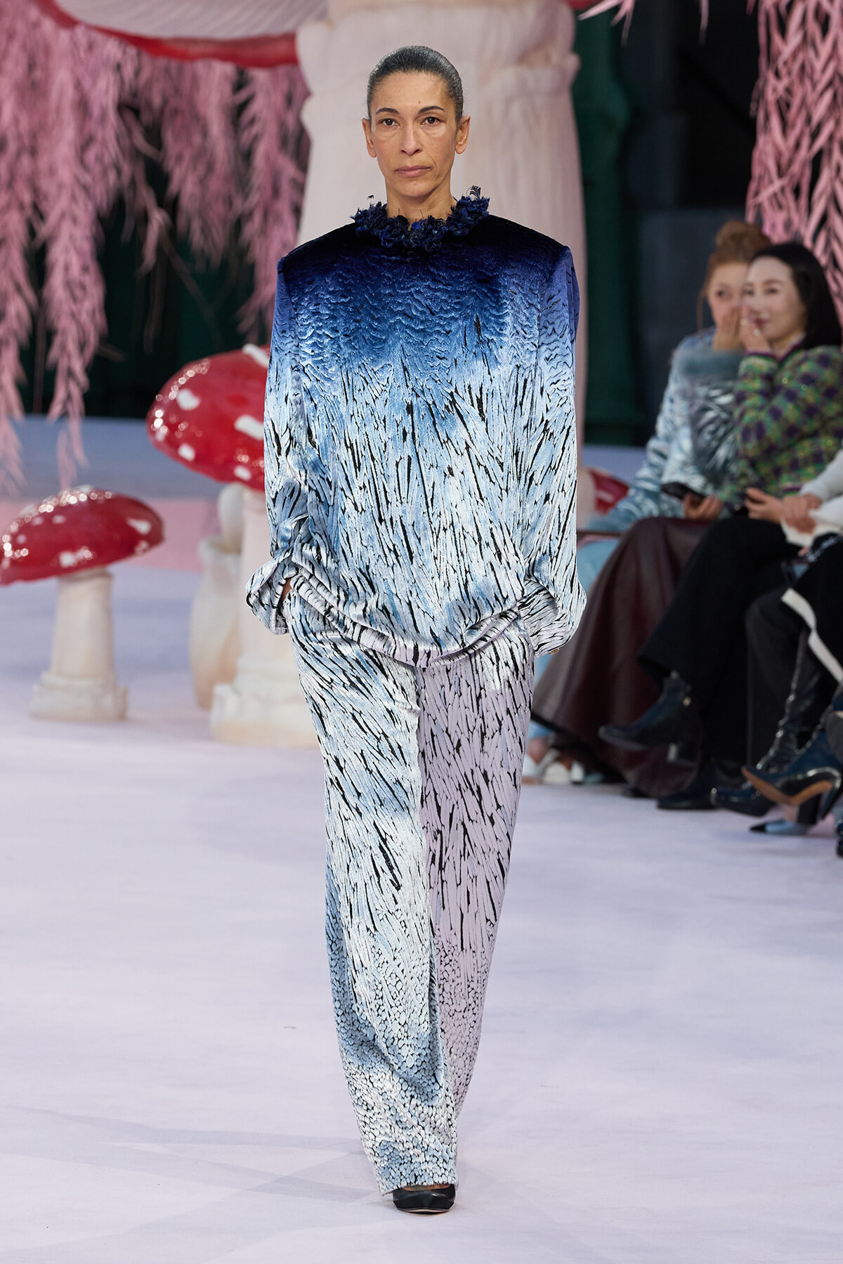 Model walks a runway in a gradient blue to white textured jumpsuit with a dark blue ruffled neckline; audience sits in the background.