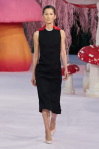 Model walking a runway in a sleeveless black midi dress with a red neck scarf and pale pink heels, against a whimsical backdrop.