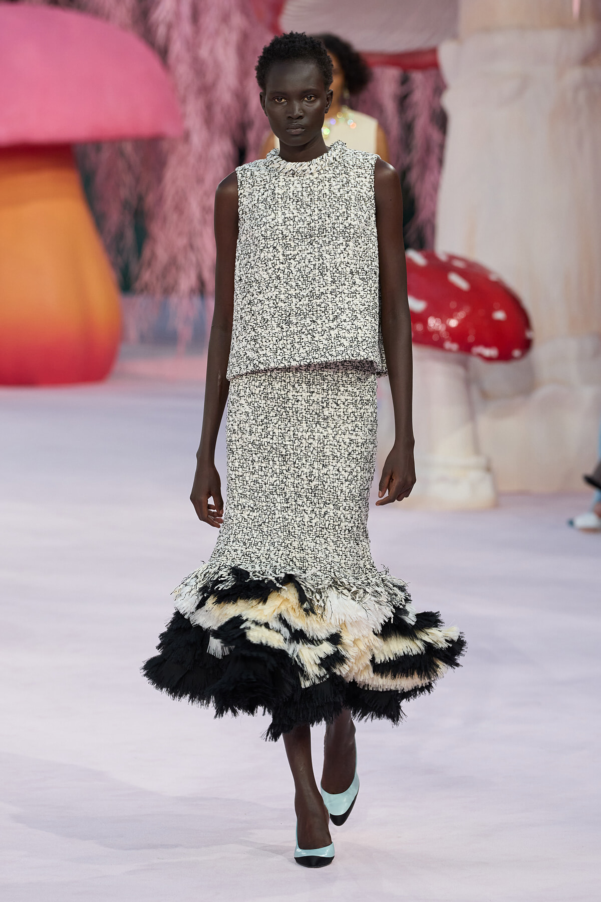 Model on a runway wearing a sleeveless speckled two-piece set with a dramatic black-and-white feathered hem and mint shoes