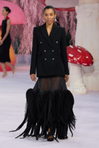 Model in a black double-breasted blazer with green gems and a sheer feathered-hem skirt on the runway.