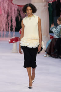 Model on a runway wearing an ivory sleeveless top with a fringe hem and layered cream feathered skirt over black cropped pants, white heels.