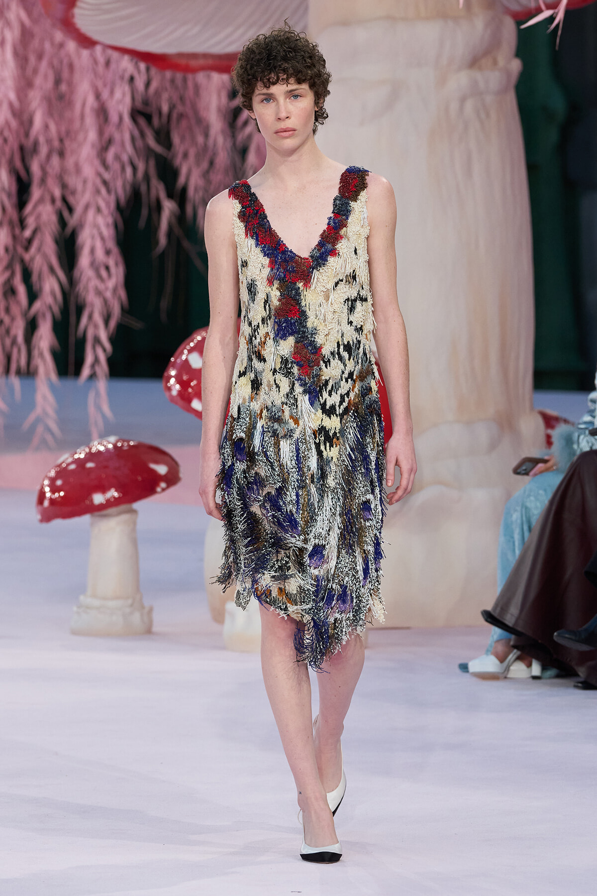 Female model walking a runway in a fringed, multicolored dress with red, blue, and cream tones.