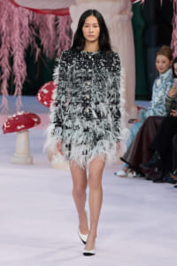 Model walking a runway in a black tweed mini dress densely trimmed with white feathers, paired with white-and-black heels.