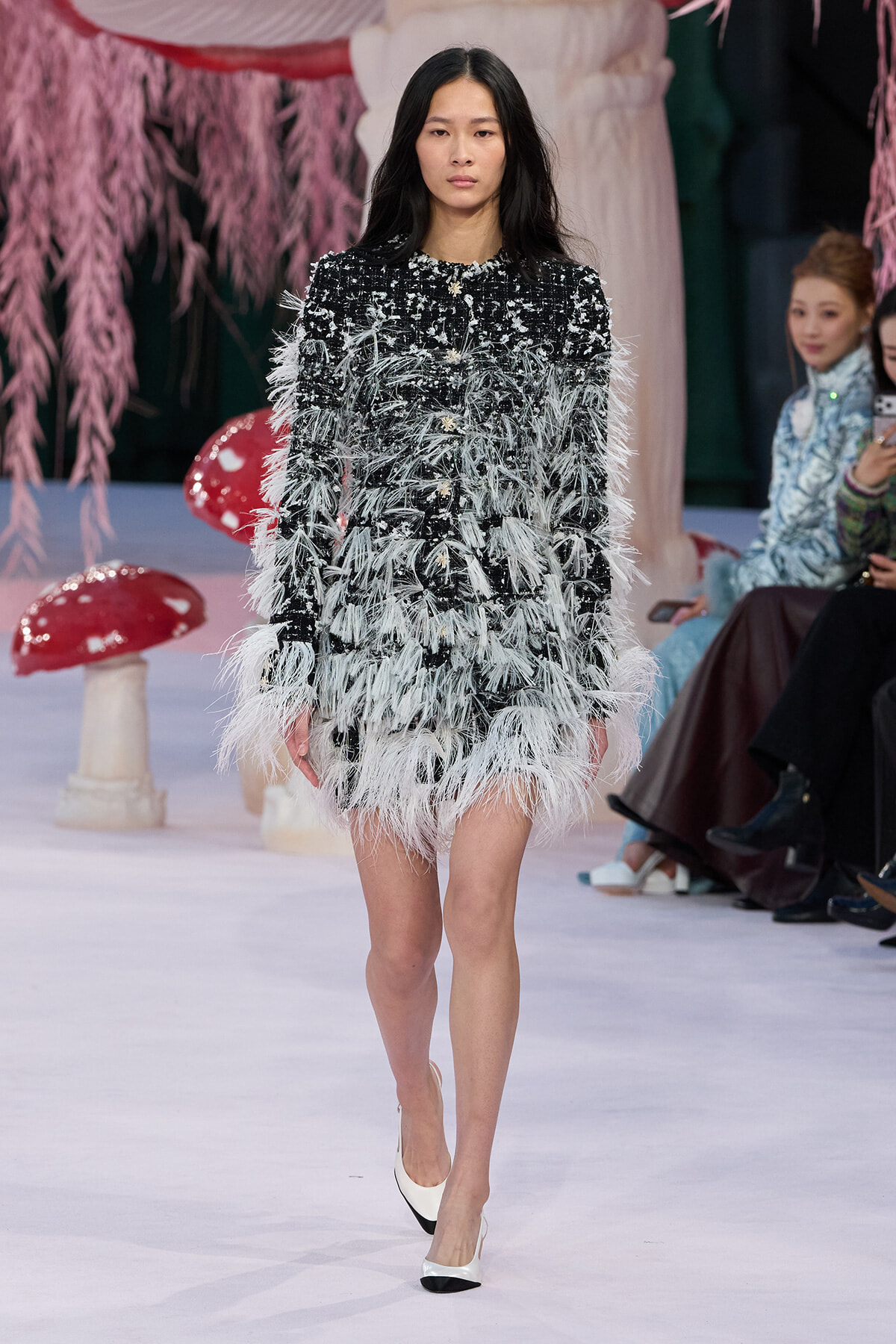 Model walking a runway in a black tweed mini dress densely trimmed with white feathers, paired with white-and-black heels.