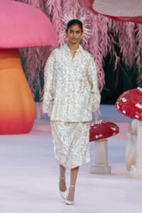 Model walks the runway in a white sequined suit with a spiky starburst headpiece, pastel set design in background.