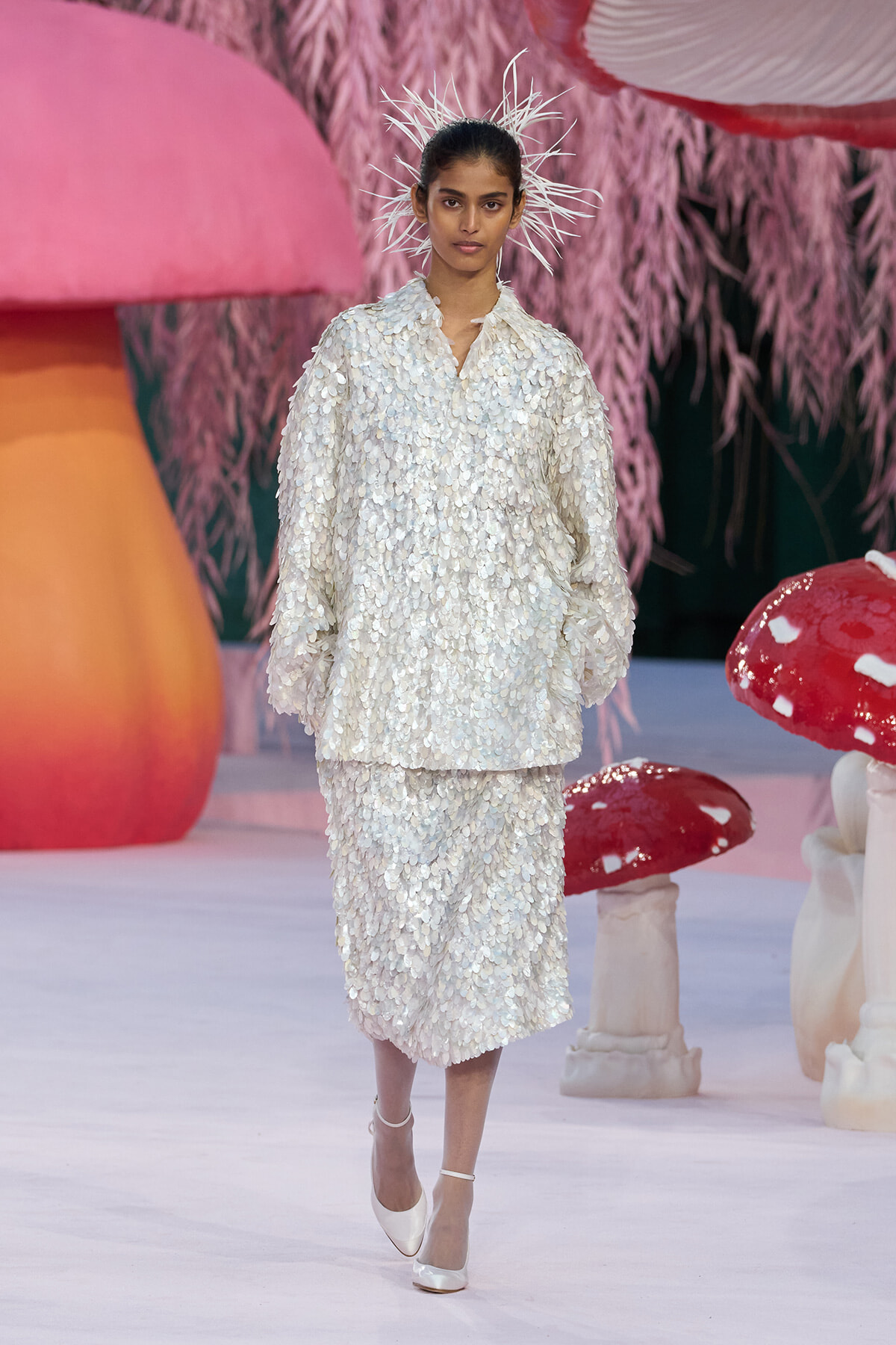 Model walks the runway in a white sequined suit with a spiky starburst headpiece, pastel set design in background.