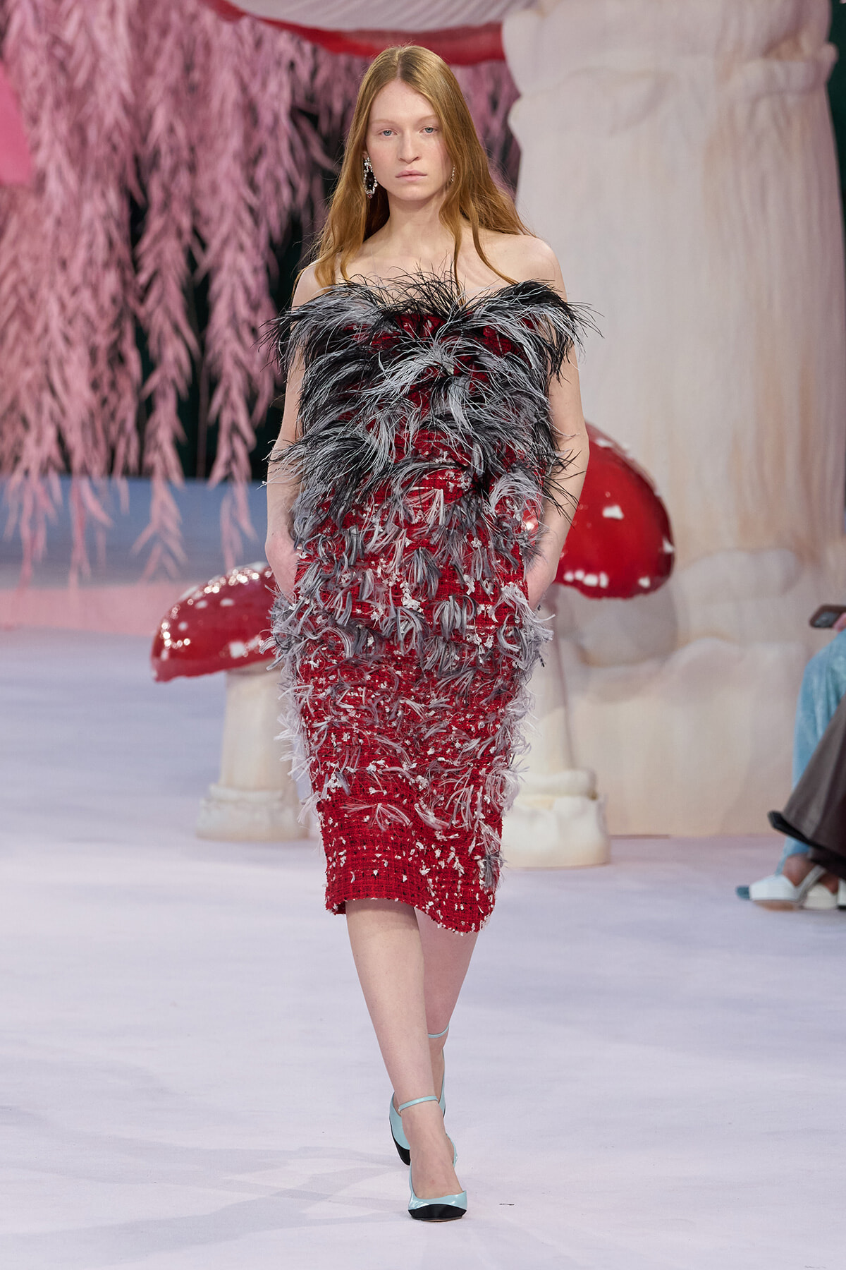 Model walks the runway in a red sequined dress with black and grey feather accents, paired with light blue heels.