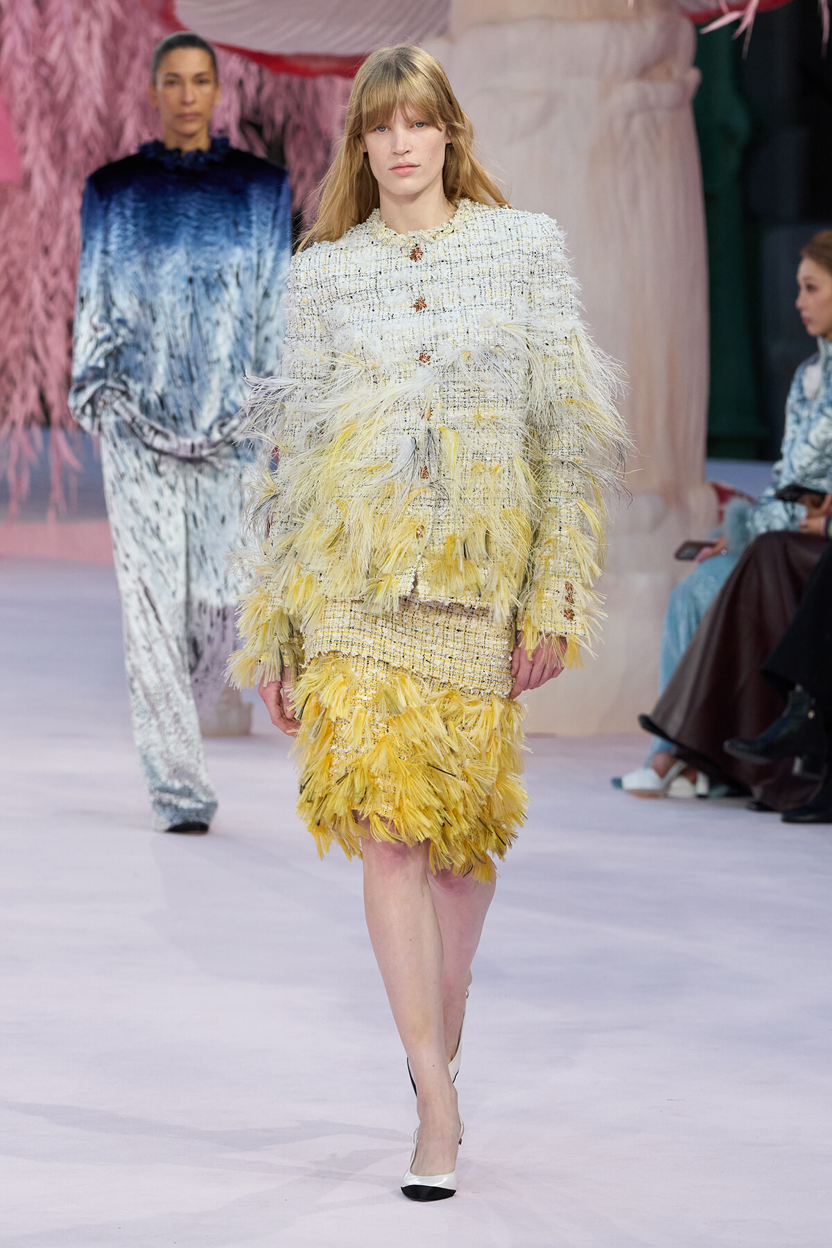 Model walking a fashion show in a cream tweed dress with yellow feathered fringe and matching skirt; pastel runway setting in background.