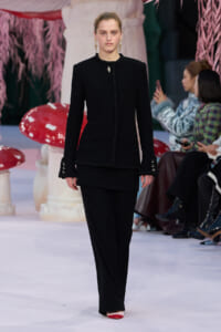Female model walks a fashion runway in a black fringed-texture suit with matching wide-leg pants, accessorized with gold earrings and a pendant necklace.