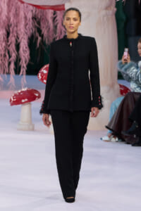 Model on a runway wearing a black tailored jacket with fringe along the front and cuffs, paired with matching black pants.