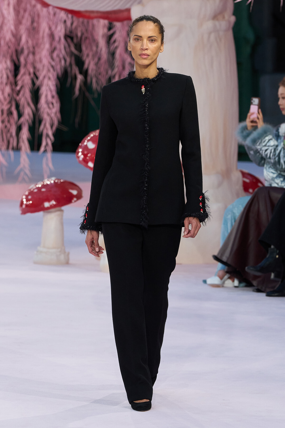 Model on a runway wearing a black tailored jacket with fringe along the front and cuffs, paired with matching black pants.
