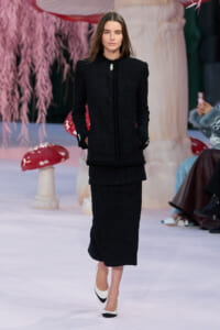 Model on a runway wearing a black textured bouclé suit with a long skirt and mint-green accents, posing with hands in pockets.