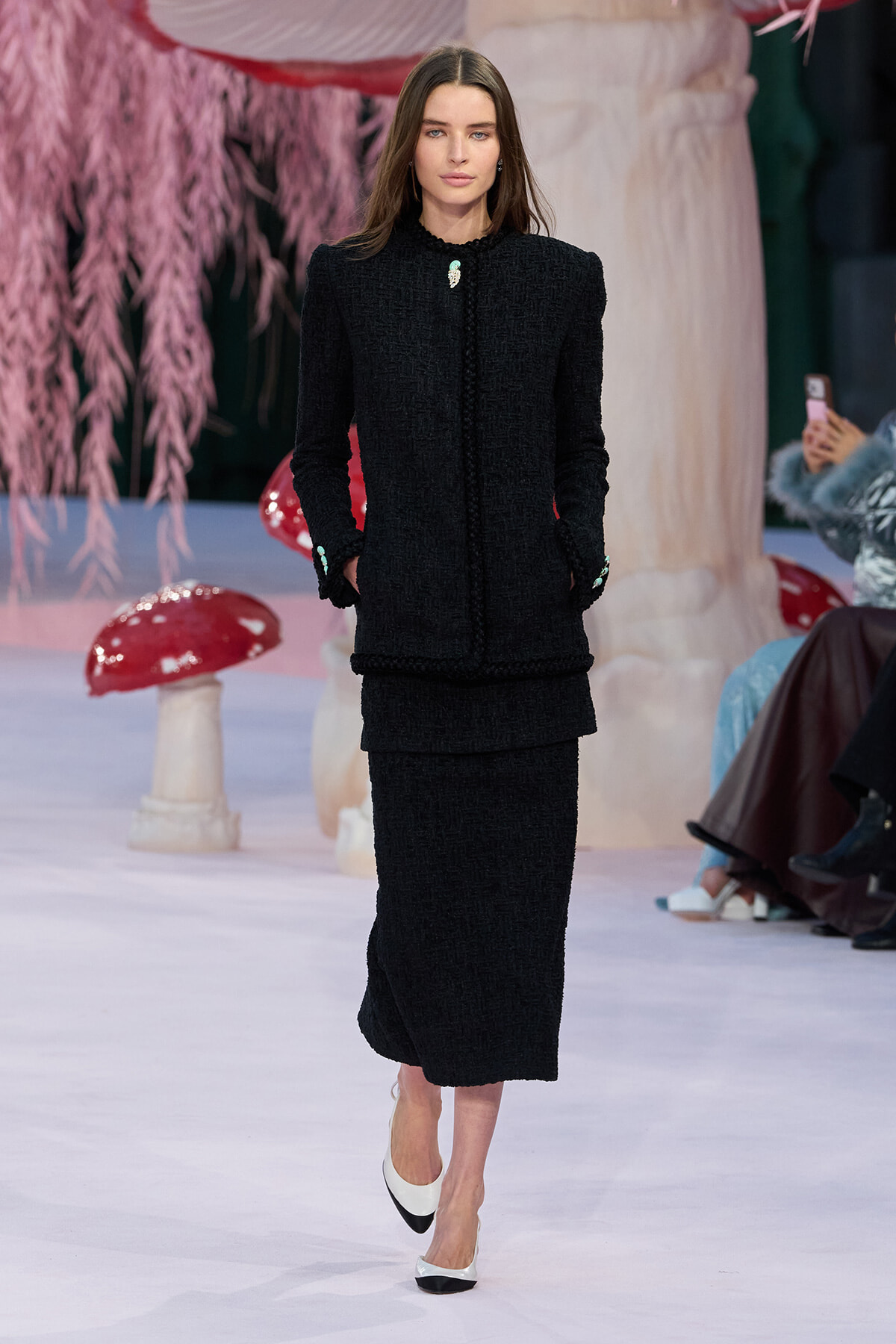Model on a runway wearing a black textured bouclé suit with a long skirt and mint-green accents, posing with hands in pockets.