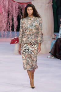 Model walks the runway wearing a textured, multicolor tweed suit with matching skirt and beige heels, fashion show scene.
