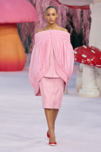 Model walking a runway in a pink off-the-shoulder, voluminous cape over a matching pink textured dress, paired with red heels.