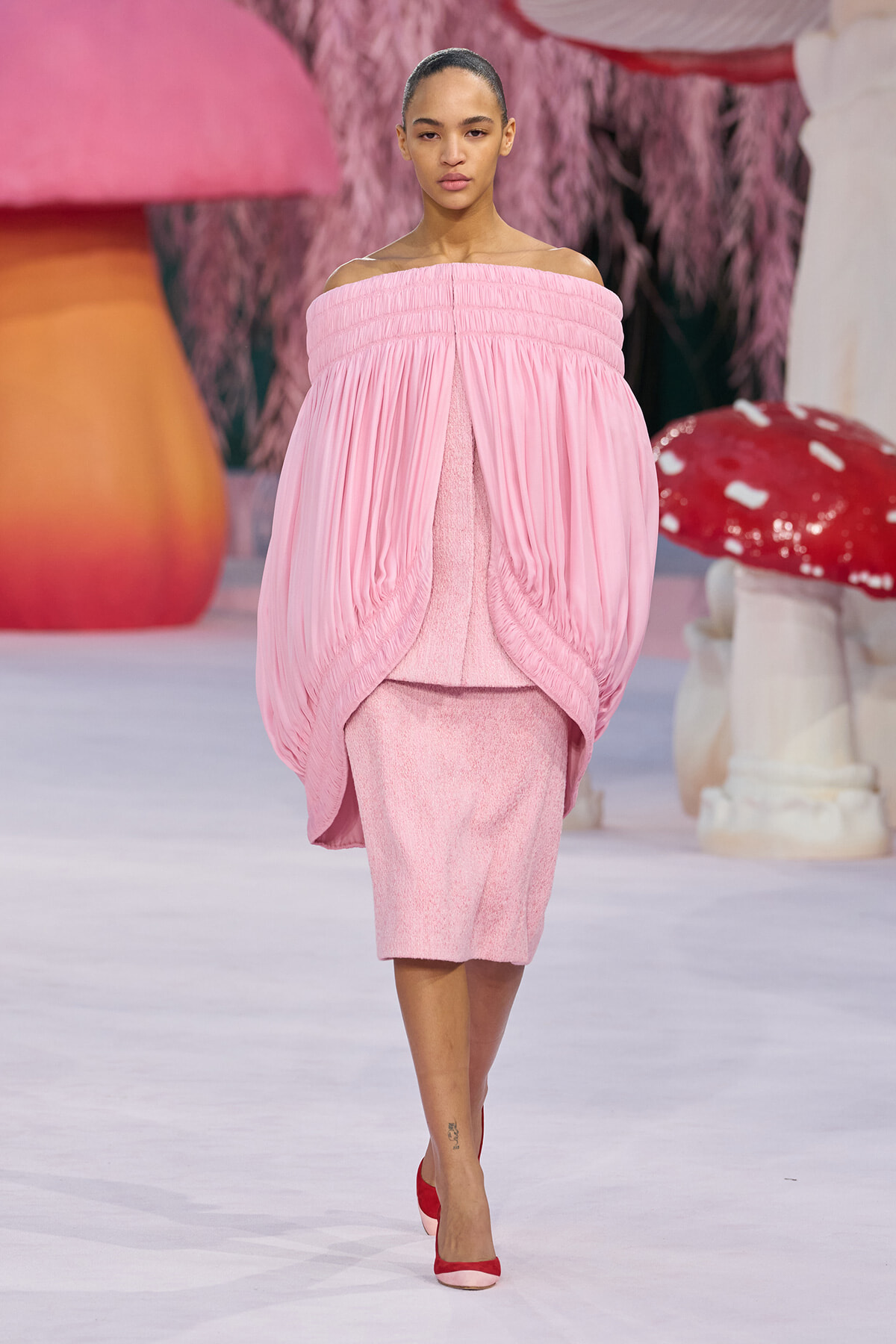 Model walking a runway in a pink off-the-shoulder, voluminous cape over a matching pink textured dress, paired with red heels.