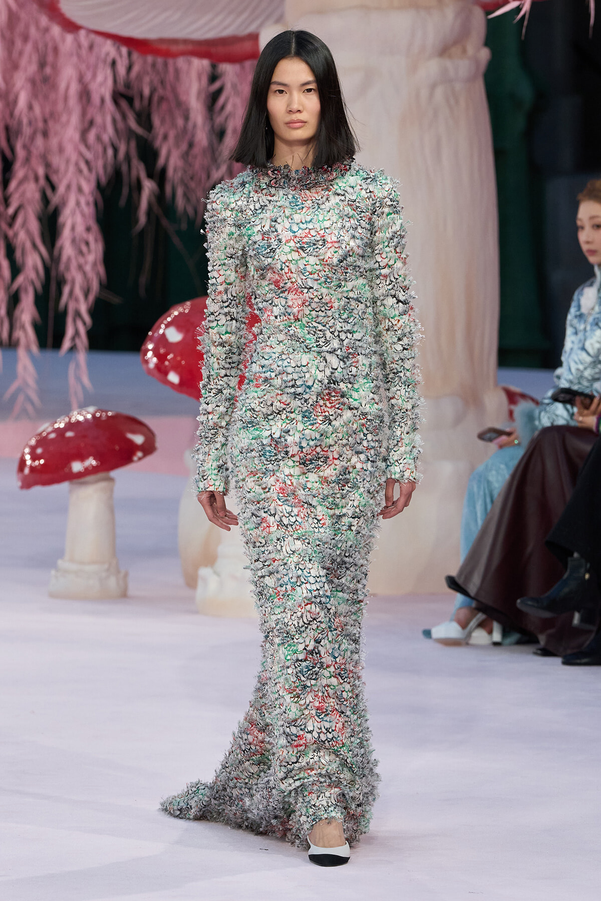 Model walks the runway in a full-length, textured multicolor fringe gown with black hair and white shoes, set against whimsical pink decor and oversized mushrooms.