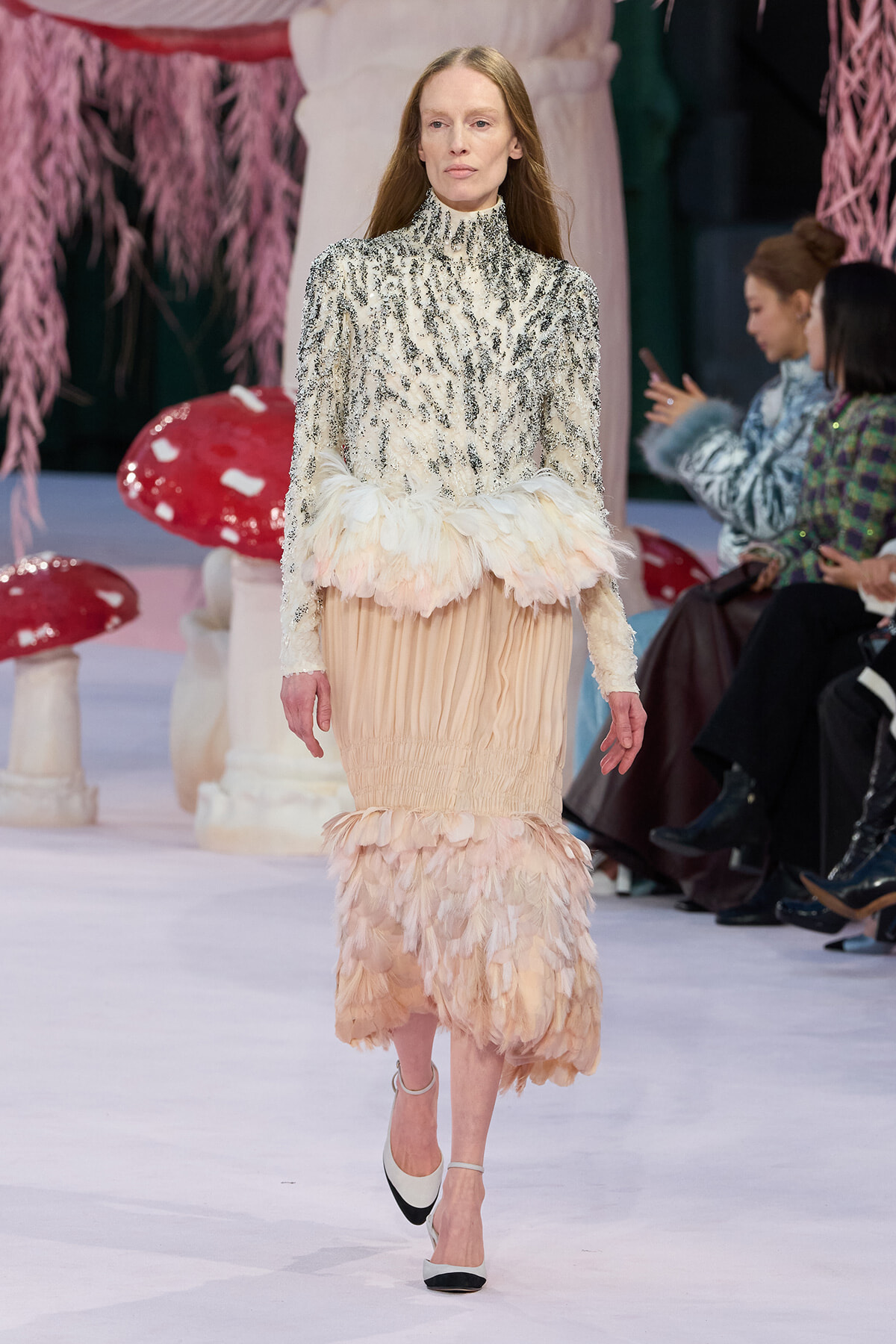 Model on a runway wearing a high-neck silver sequin blouse and a pale peach feather-trimmed skirt with white-and-black heels