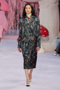 Runway model walks in a knee-length, textured dark tweed coat with bright green buttons on a fashion show set.