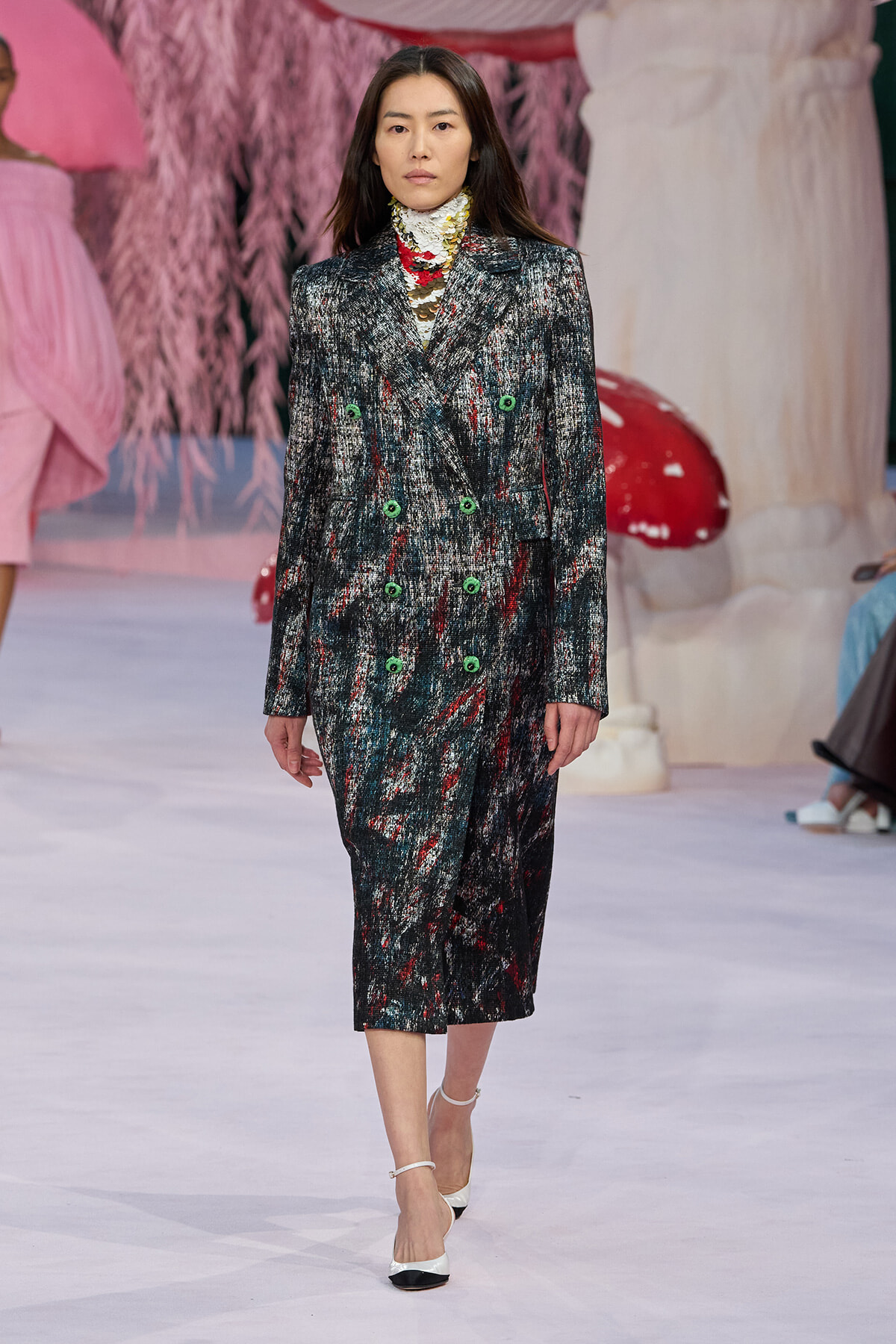 Runway model walks in a knee-length, textured dark tweed coat with bright green buttons on a fashion show set.