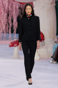 Female model walking runway in a black tailored suit with colorful floral collar and cuffs, pairing black pants with black heels amid pastel stage décor.