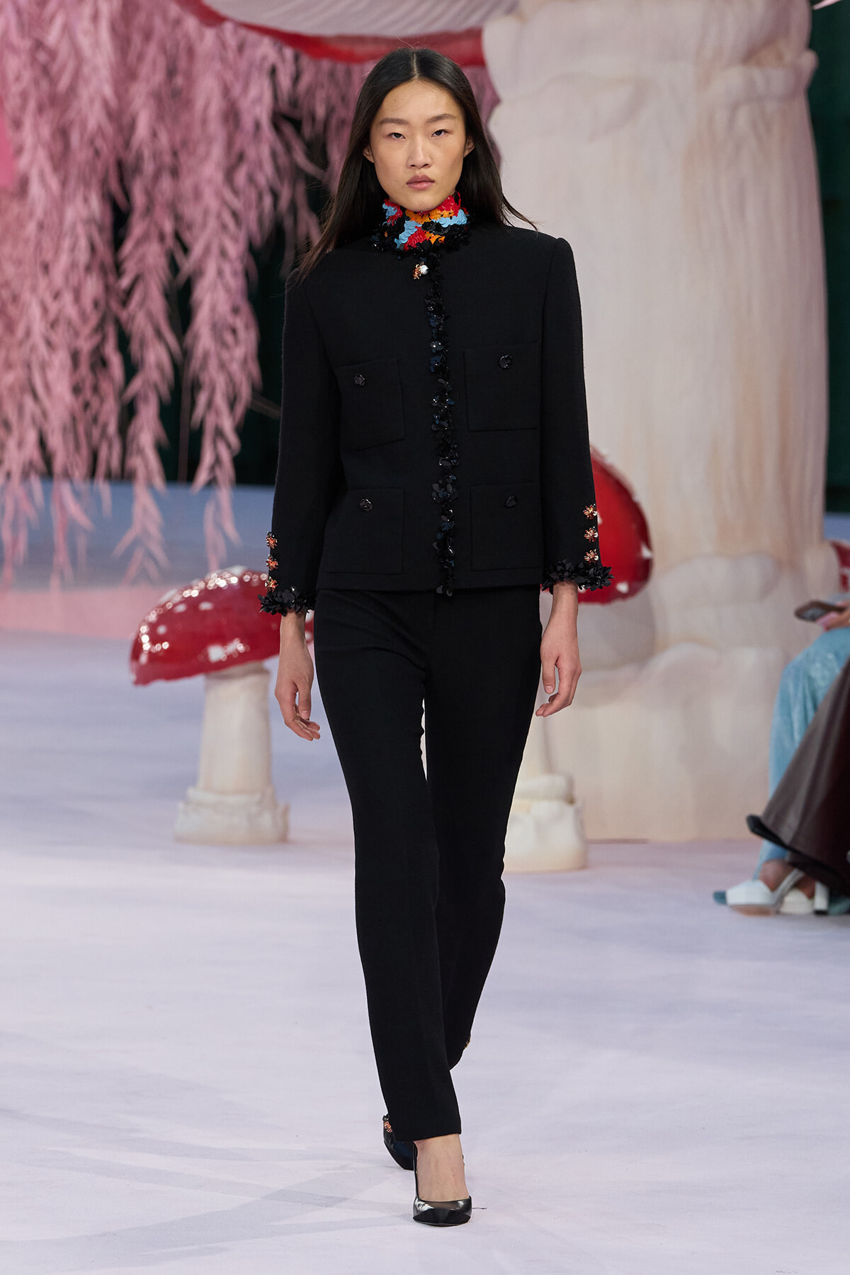 Female model walking runway in a black tailored suit with colorful floral collar and cuffs, pairing black pants with black heels amid pastel stage décor.