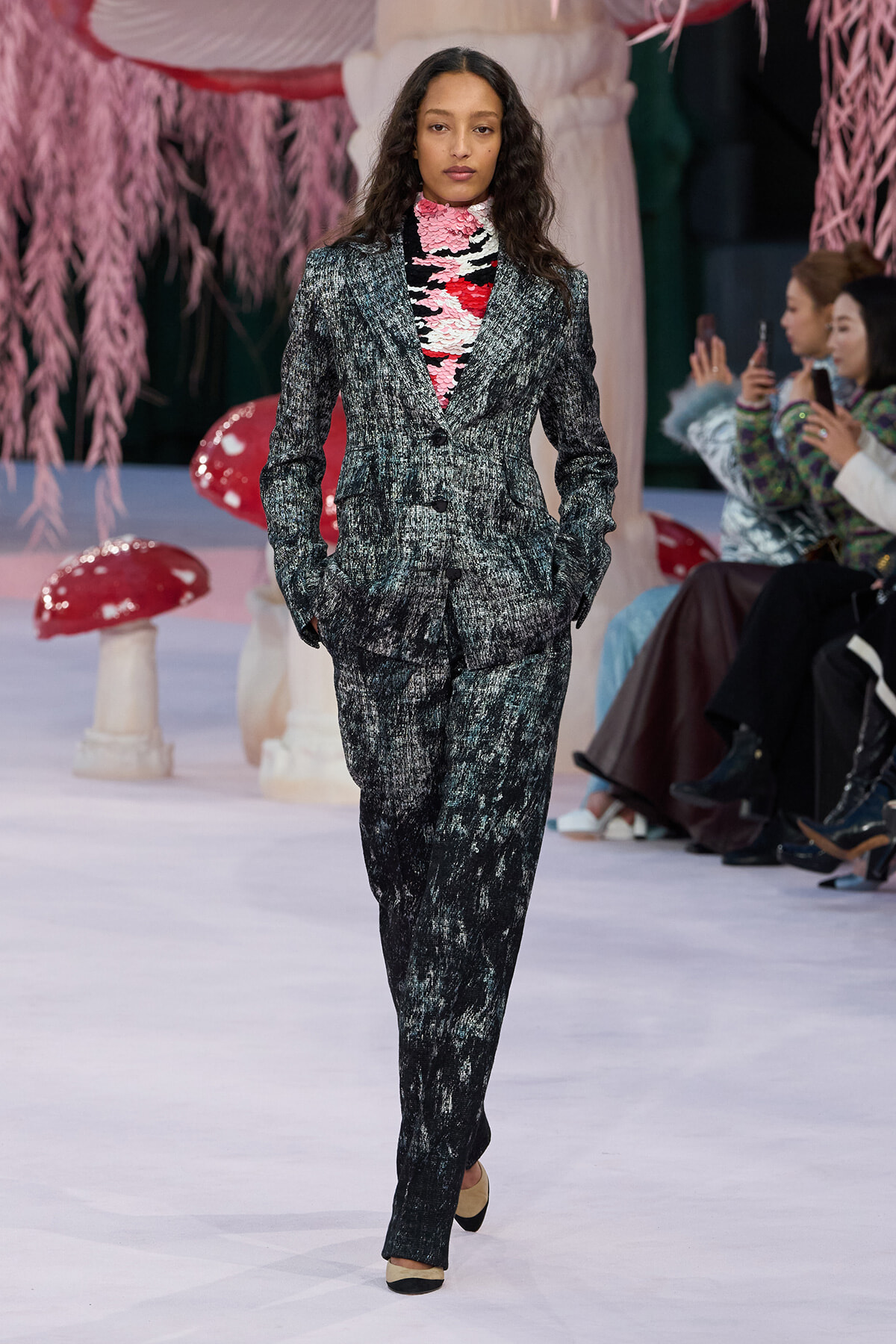 Female model walking a runway in a metallic textured gray suit with a pink floral turtleneck visible at the neck, hands in pockets.
