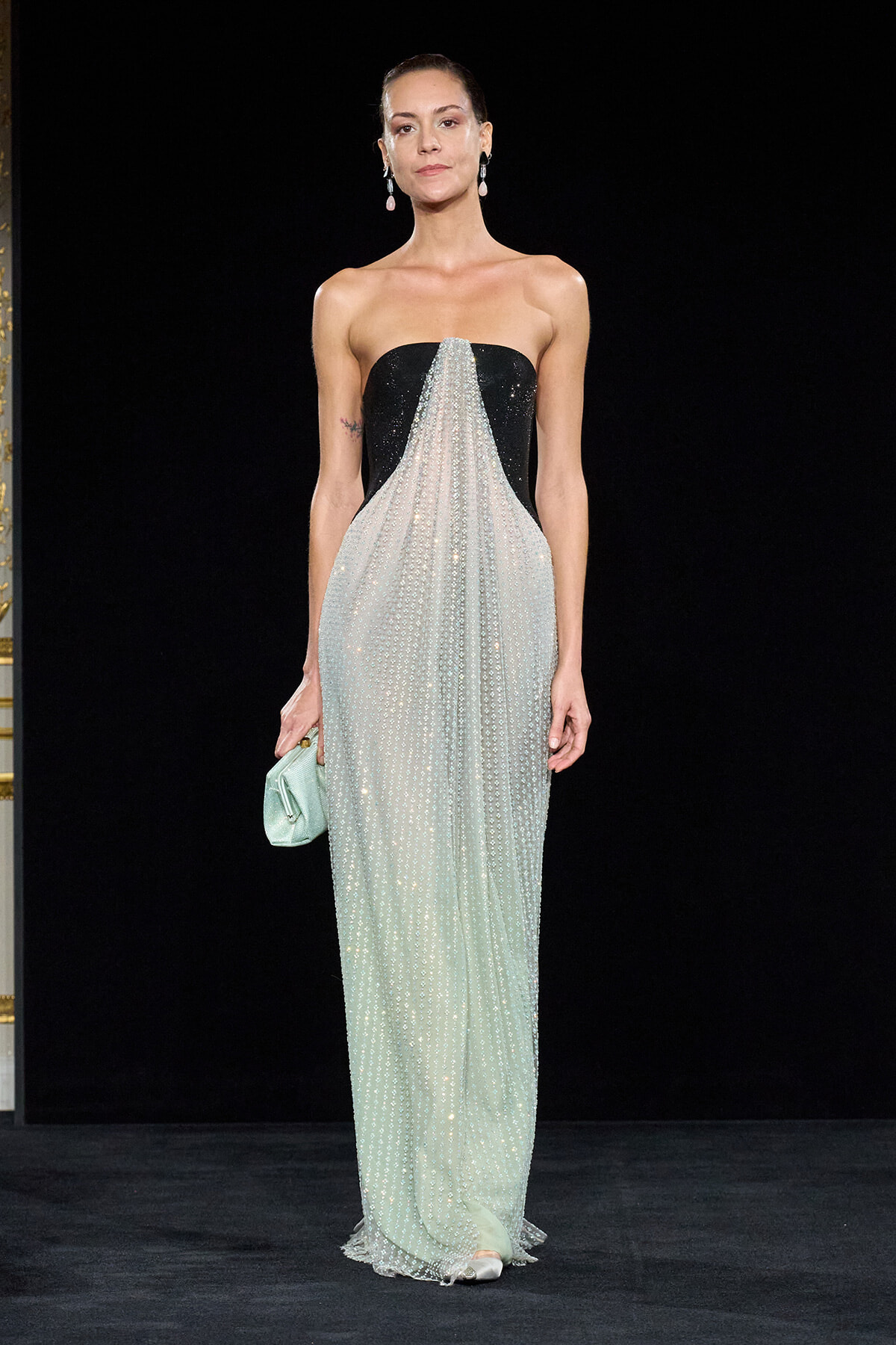 Model on a runway wearing a strapless black gown with a sparkling mint-beaded overlay and a mint clutch.