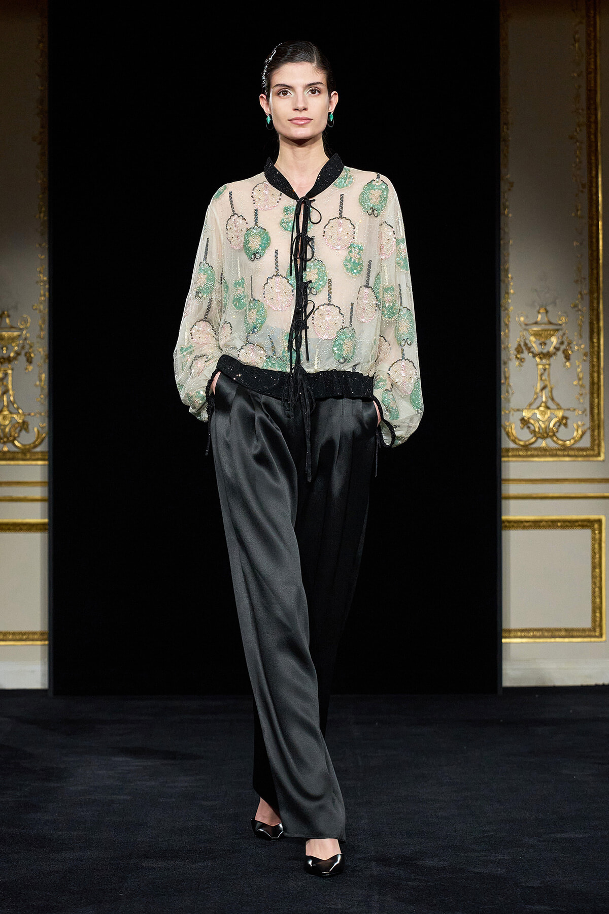 Model walking a runway in a sheer floral blouse with green and pink blossoms, black collar and waist tie, paired with black satin wide-leg pants.