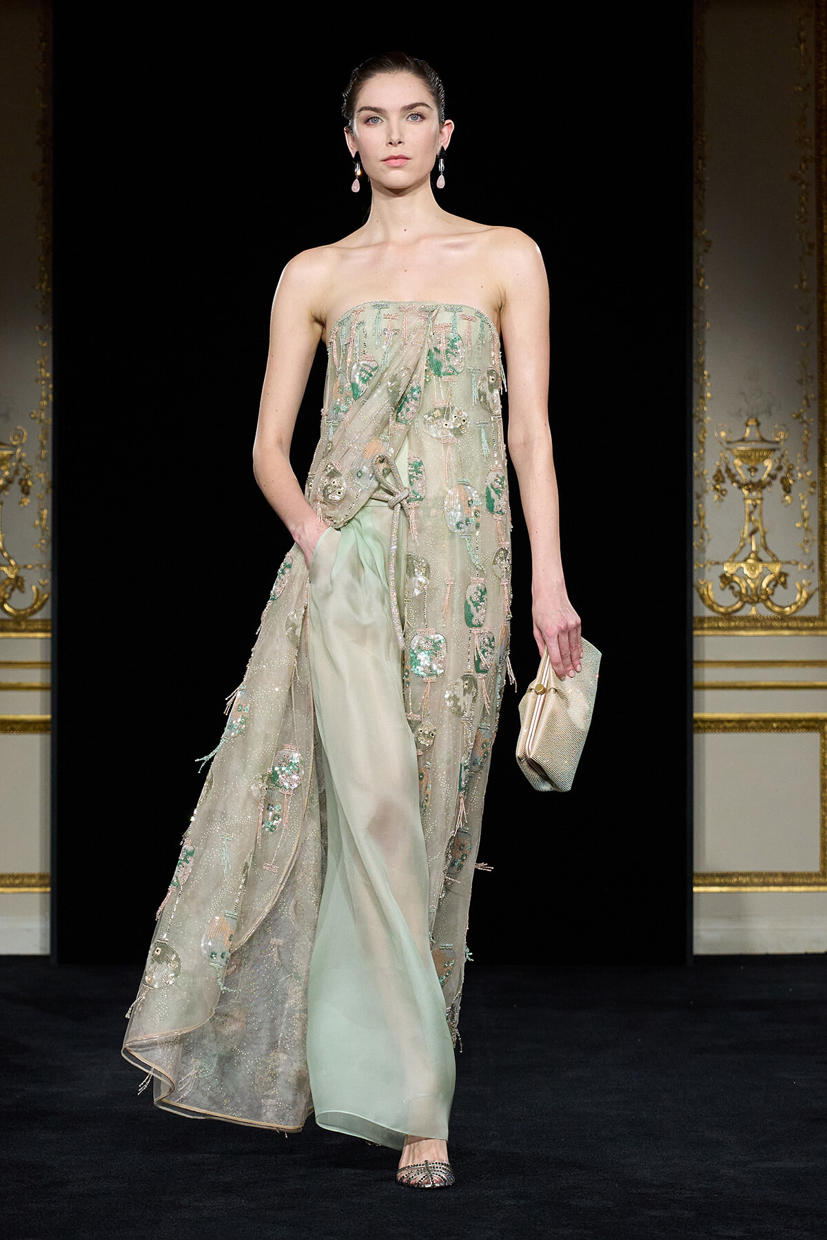 Model on a runway wearing a strapless, pale green embellished gown with a sheer overlay and matching clutch.
