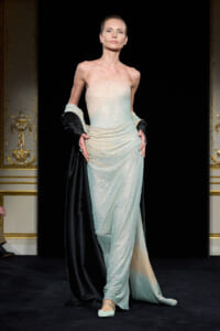 Model on a runway wearing a strapless, sparkling mint gown with a black cape draped over the shoulders.