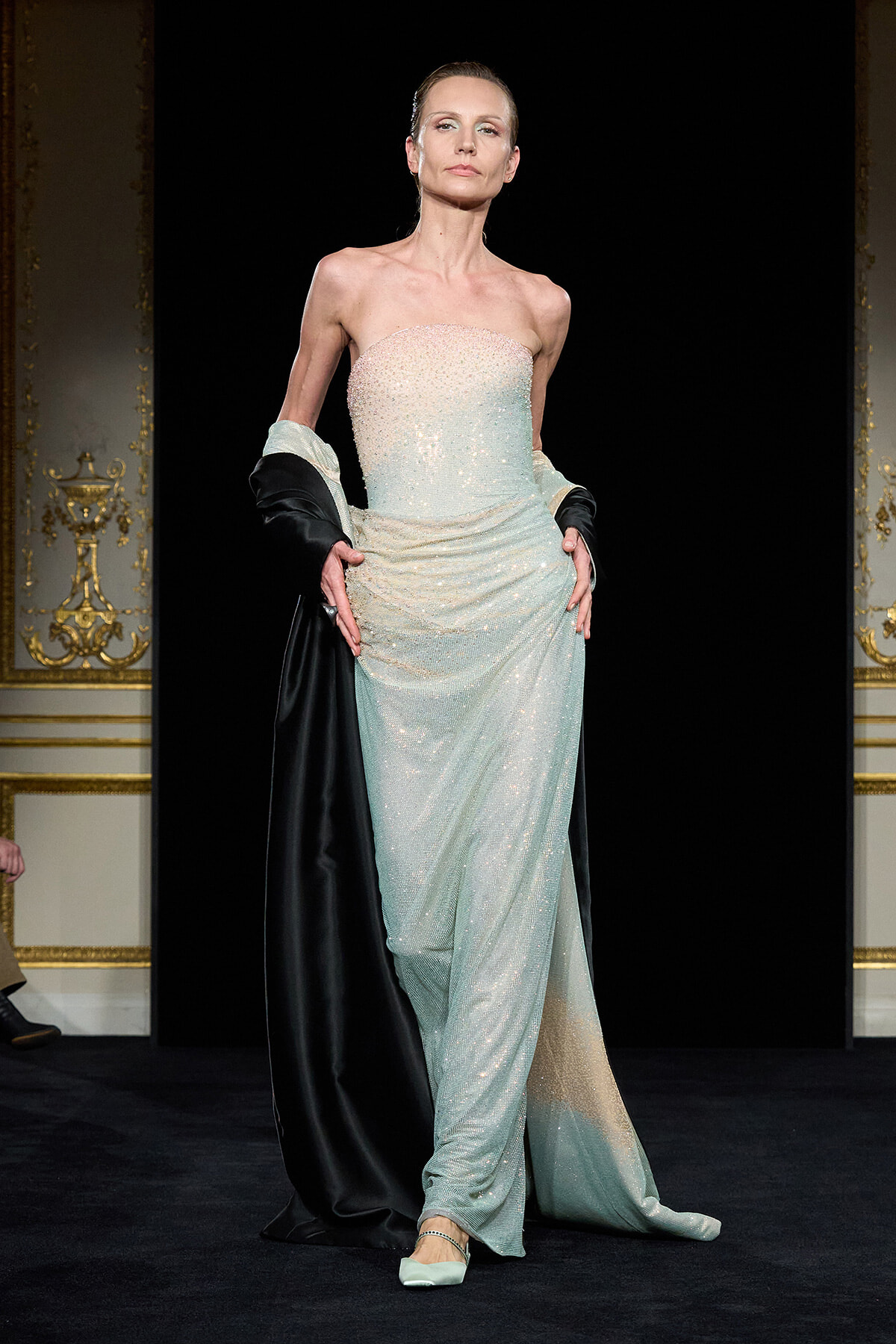 Model on a runway wearing a strapless, sparkling mint gown with a black cape draped over the shoulders.